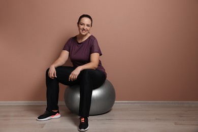 Happy overweight woman sitting on fitness ball near pale pink wall indoors. Space for text