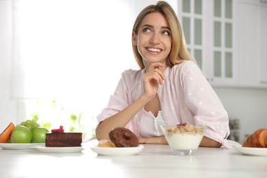 Woman choosing between sweets and healthy food at white table in kitchen