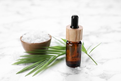 Bottle of essential oil, sea salt and green twig on white marble table, closeup