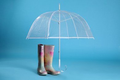Open transparent umbrella and rubber boots on light blue background