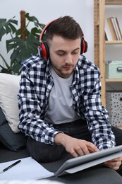 Photo of Young man with modern tablet and headphones studying on sofa at home. Distance learning