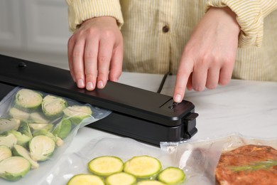 Woman using sealer for vacuum packing with plastic bag of zucchini at white table, closeup