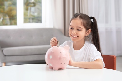 Little girl putting money into piggy bank at table indoors