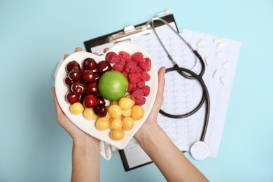 Female doctor holding plate with fresh fruits over color background, top view. Cardiac diet
