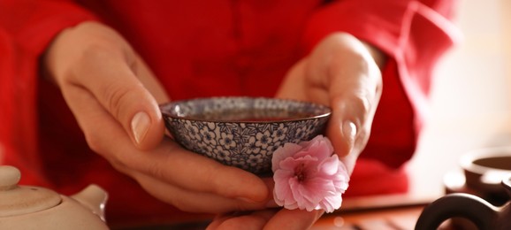 Guest holding cup of freshly brewed tea and sakura flower during traditional ceremony at table indoors, closeup. Banner design
