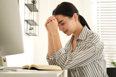 Beautiful young woman praying over Bible at desk
