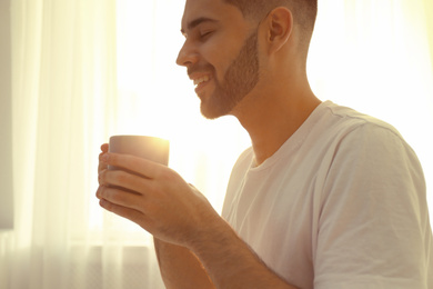 Young man with cup of coffee near window at home, closeup. Lazy morning