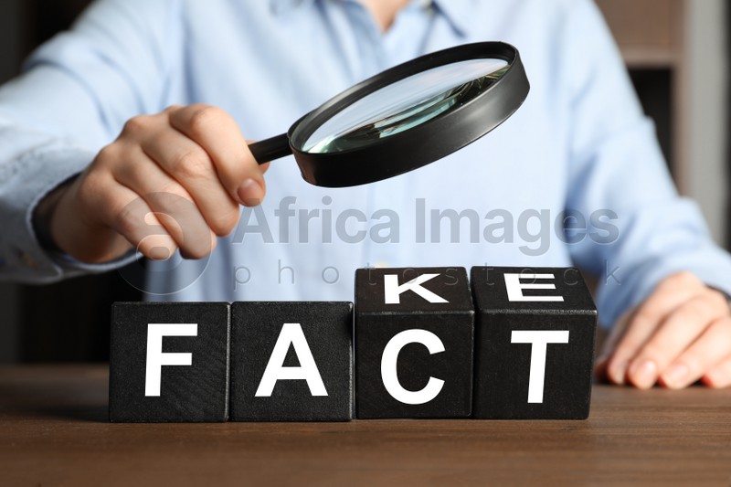 Woman holding magnifying glass above words FAKE and FACT made with black cubes at wooden table, closeup