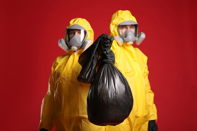 Man and woman in chemical protective suits holding trash bag on red background. Virus research