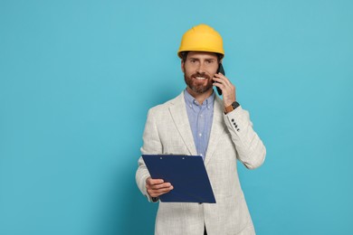 Professional engineer in hard hat with clipboard talking on phone against light blue background
