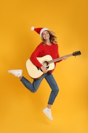 Young woman in Santa hat jumping with acoustic guitar on yellow background. Christmas music