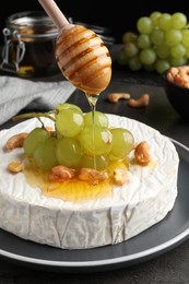 Pouring honey onto brie cheese served with grape and cashew nuts on dark grey table, closeup