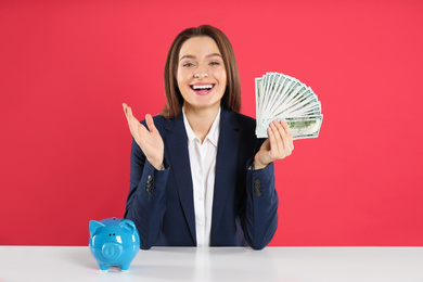 Young woman with money and piggy bank at table on crimson background