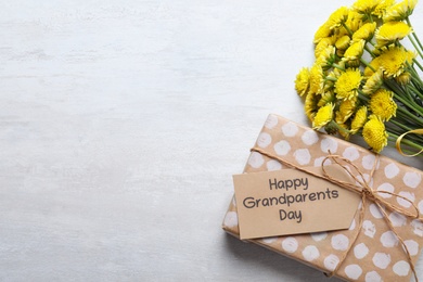 Beautiful yellow flowers, gift box and tag with phrase Happy Grandparents Day on light background, flat lay. Space for text