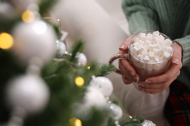 Woman with cup of delicious hot drink near Christmas tree indoors, closeup. Space for text