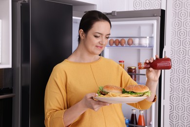 Overweight woman with ketchup and burgers near fridge in kitchen