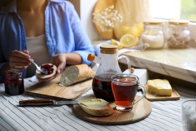 Woman spreading jam onto bread at table indoors, focus on aromatic tea