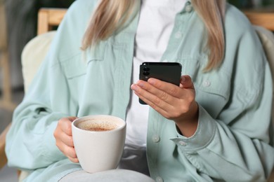 Woman with smartphone and cup of coffee indoors, closeup