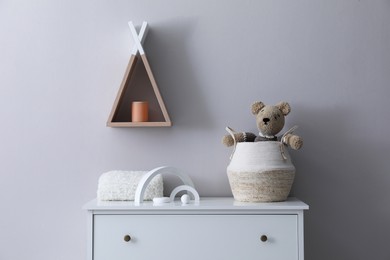 Child's toys, wicker basket and plaid on chest of drawers near light grey wall indoors