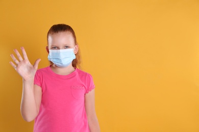 Little girl in protective mask showing hello gesture on yellow background, space for text. Keeping social distance during coronavirus pandemic
