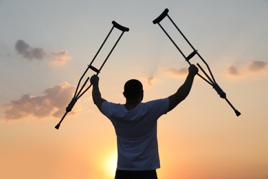 Man raising hands with underarm crutches up to sky outdoors at sunset, back view. Healing miracle