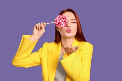 Photo of Stylish redhead woman covering eye with lollipop and blowing kiss on purple background