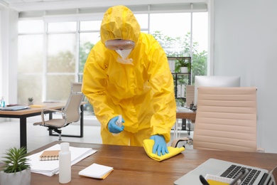 Janitor in protective suit disinfecting office furniture to prevent spreading of COVID-19