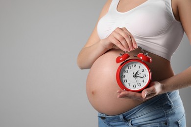 Photo of Young pregnant woman holding alarm clock near her belly on grey background, closeup and space for text. Time to give birth