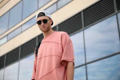 Handsome young man with stylish sunglasses and backpack near building outdoors, low angle view