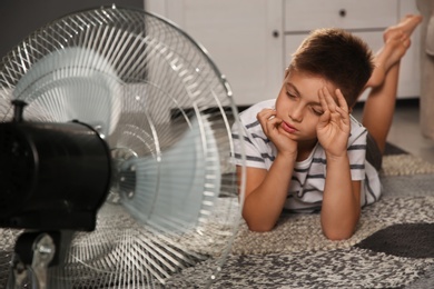 Little boy suffering from heat in front of fan at home. Summer season