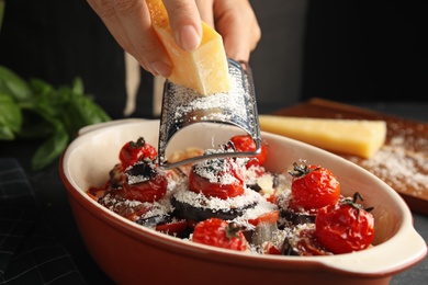 Woman grating cheese onto baked eggplant with tomatoes at table, closeup