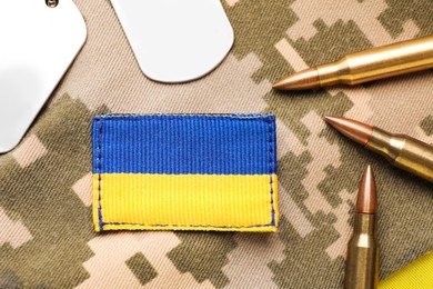 Military ID tags near Ukrainian army patch and bullets on camouflage background, flat lay