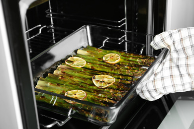 Person taking glass baking dish with cooked asparagus and lemon slices from oven, closeup
