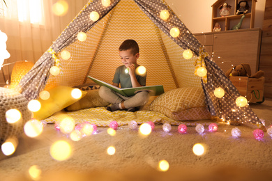 Photo of Little boy with flashlight reading book in play tent