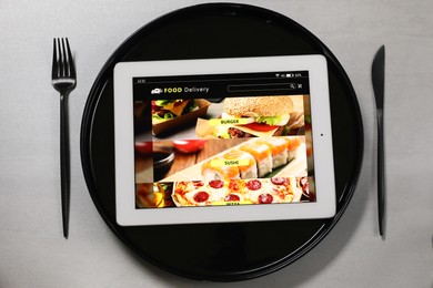 Modern tablet with open page for online food ordering, plate and cutlery on grey table, flat lay. Concept of delivery service
