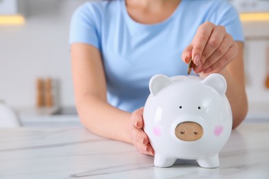 Young woman putting coin into piggy bank at table indoors, closeup and space for text. Money savings