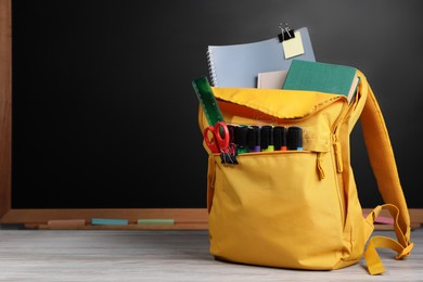 Backpack with different school stationery on white wooden table near blackboard, space for text