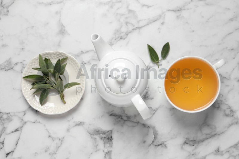 Photo of Cup of sage tea, green leaves and teapot on white marble table, flat lay