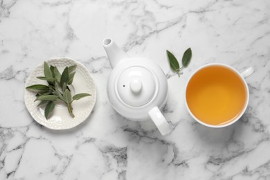 Cup of sage tea, green leaves and teapot on white marble table, flat lay