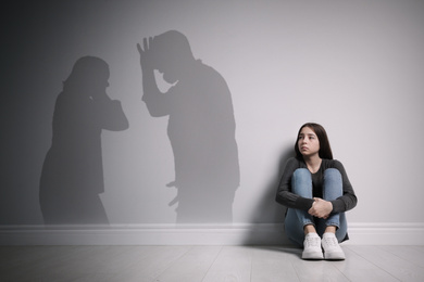 Image of Upset teenage girl sitting on floor near wall and silhouettes of arguing parents 