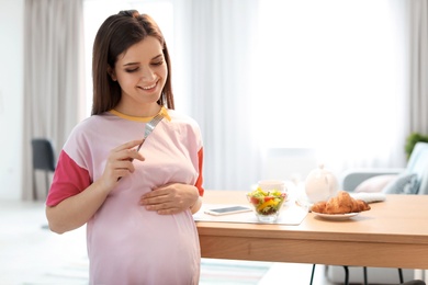 Young pregnant woman eating vegetable salad near table in kitchen