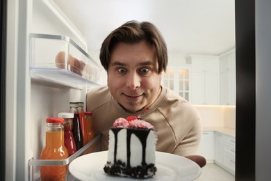 Happy overweight man taking cake out of refrigerator in kitchen, view from inside