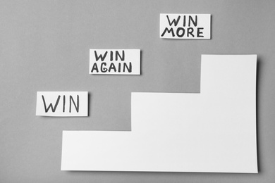 Cards with words WIN, WIN AGAIN, WIN MORE and paper cutout stairs on grey background, top view. Victory concept