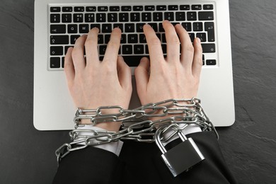 Internet addiction. Man typing on laptop with chained hands at black table, top view