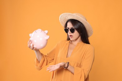 Photo of Confused young woman in stylish sunglasses and straw hat with piggy bank on orange background