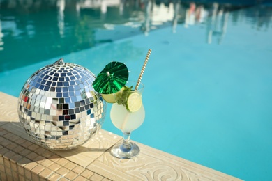 Tasty refreshing cocktail and shiny disco ball on edge of swimming pool. Party items