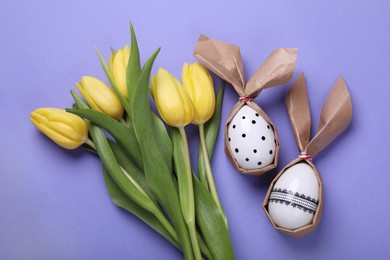 Photo of Easter bunnies made of craft paper and eggs near beautiful tulips on purple background, flat lay
