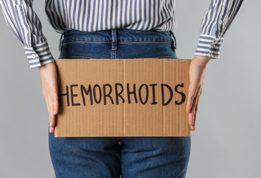 Woman holding carton sign with word HEMORRHOIDS on light grey background, closeup