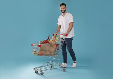 Happy man with shopping cart full of groceries on light blue background