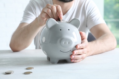 Man putting coin into piggy bank at white table indoors, closeup